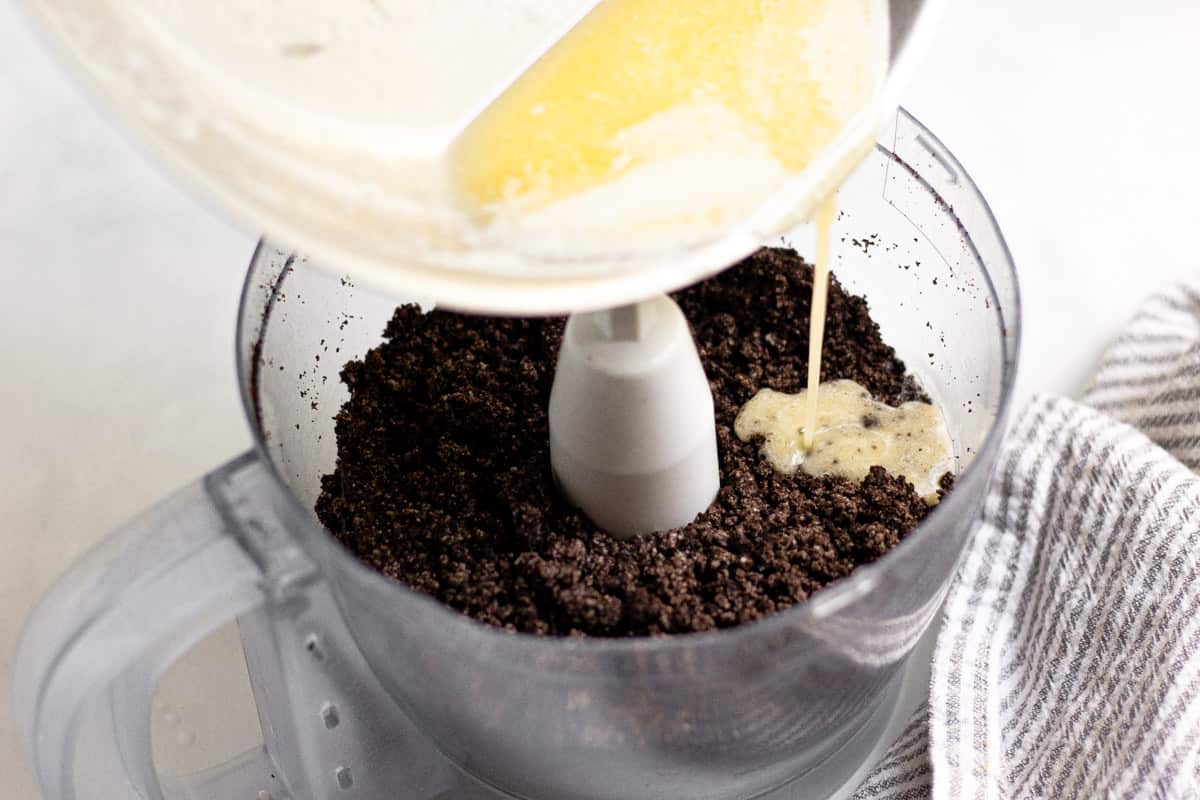 Food processor filled with oreo crumbs with butter being poured into it 