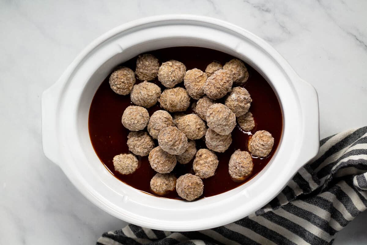 Overhead shot of an oval white crock pot insert filled with whiskey bourbon barbecue sauce and frozen meatballs