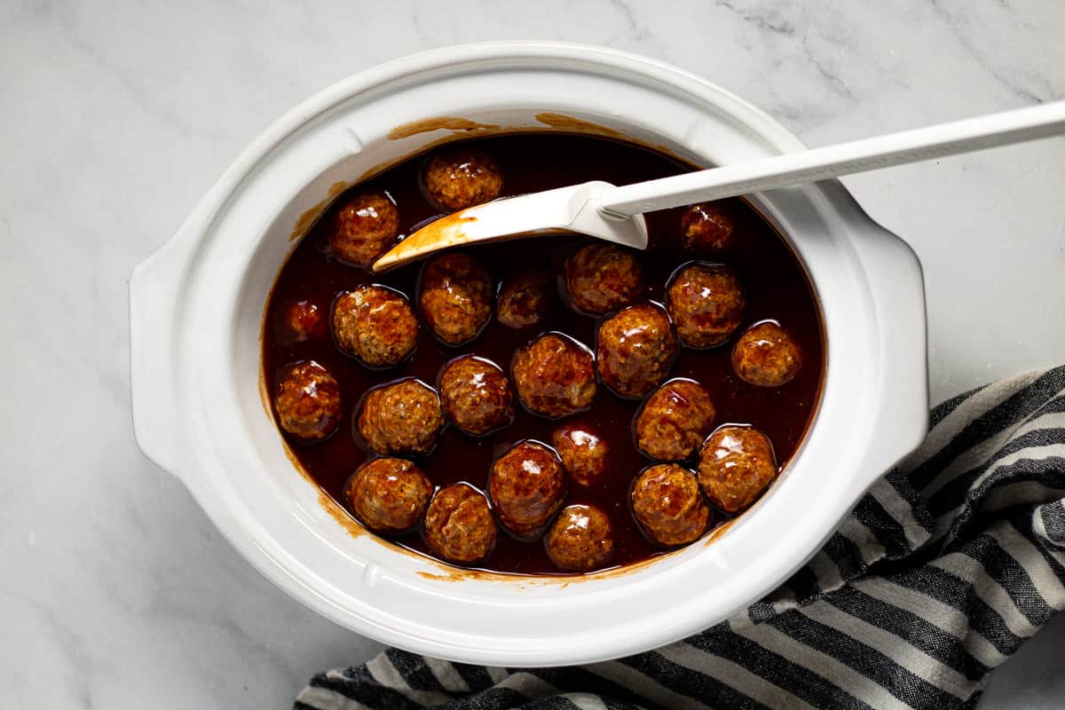 Overhead shot of an oval white crock pot insert filled with whiskey bourbon barbecue sauce and frozen meatballs