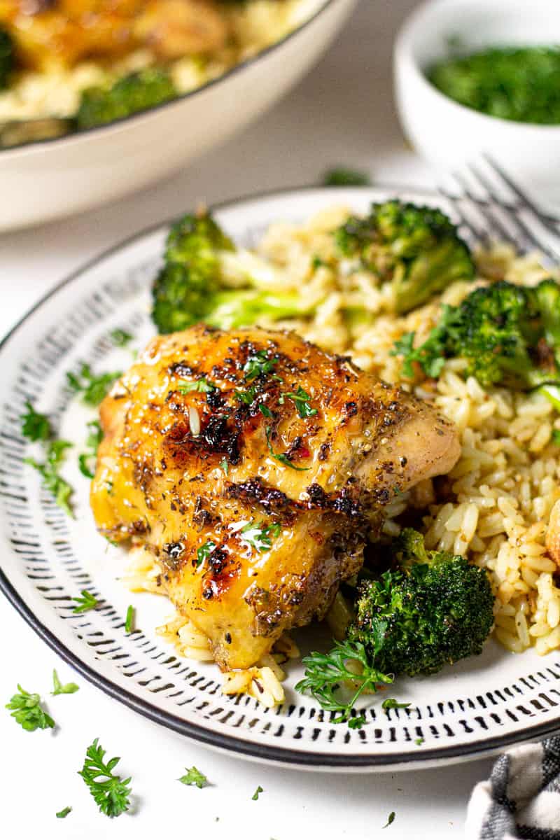 Close up shot of a roasted chicken thigh on a white plate with rice pilaf and broccoli
