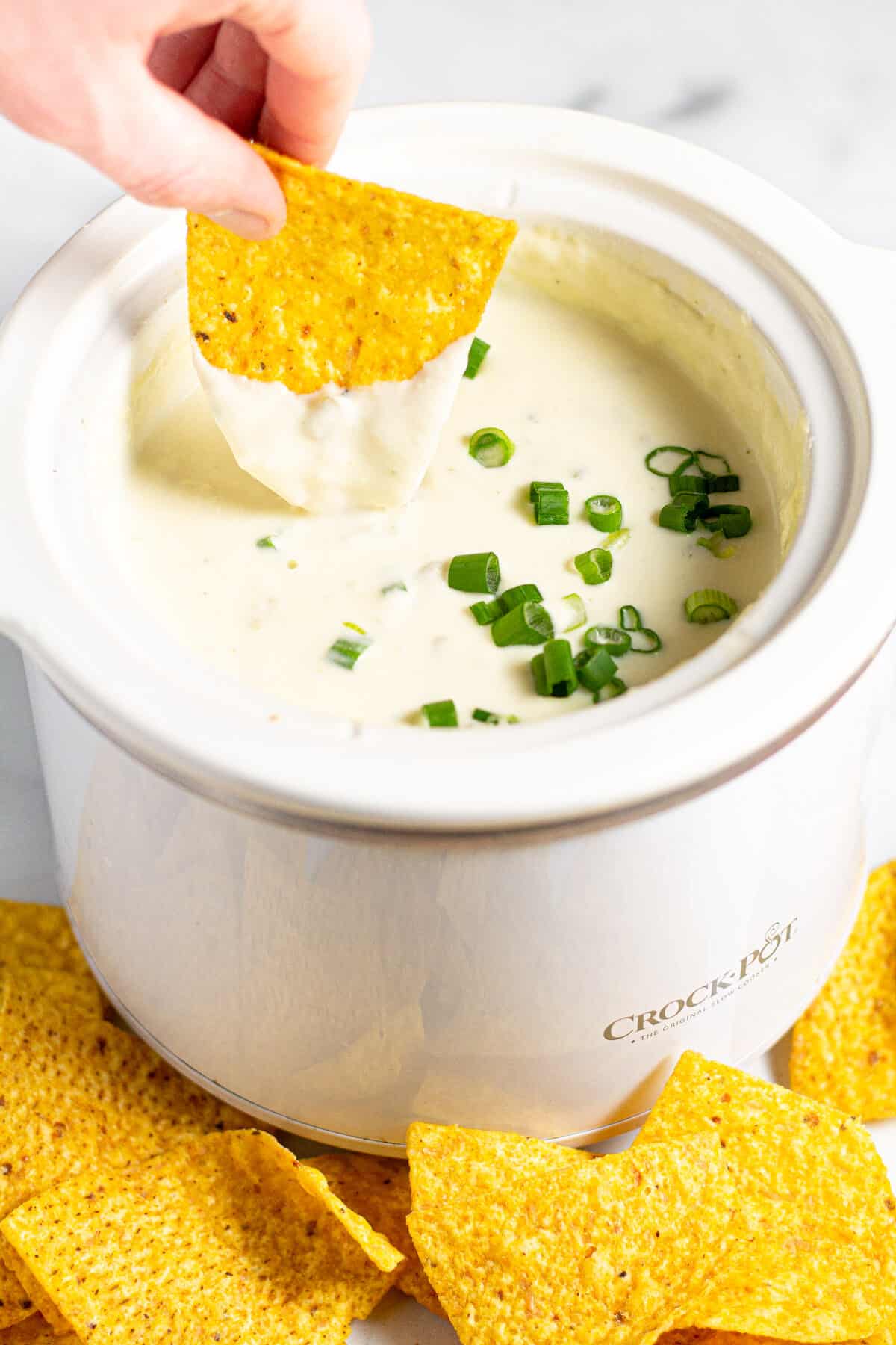 A hand holding a tortilla chip and dipping it into a crock pot of queso 