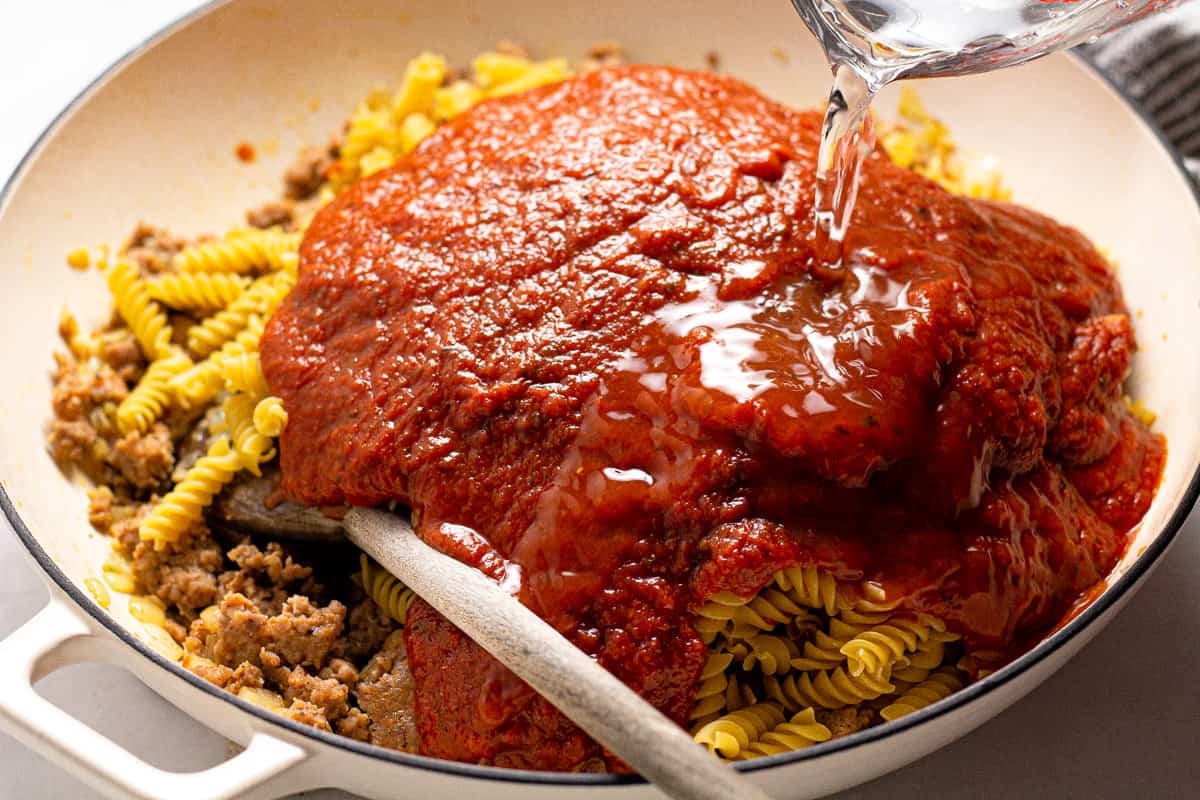 Water being poured into a large white pot filled with pasta sauce and noodles
