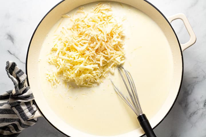 Large white pot with shredded cheese being added to a sauce 
