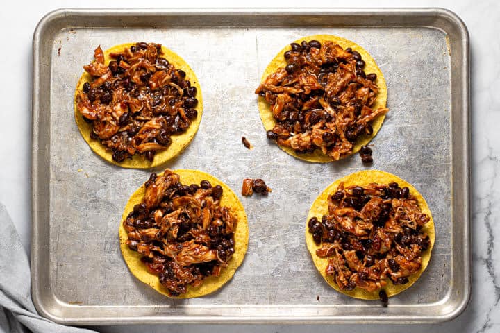 Shredded chicken and beans on top of 4 tostada shells on a baking sheet