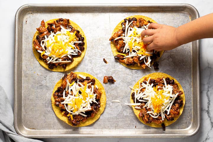 A hand adding cheese to the top of a bbq chicken tostada on a baking sheet