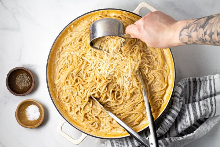 Large white pot filled with alfredo with a hand pouring Parmesan cheese over the pasta