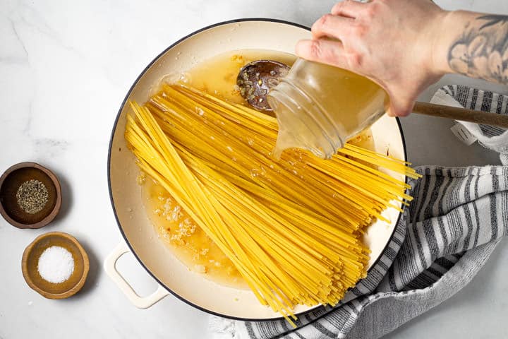 Large white pot with linguine noodles and a hand pouring broth over the noodles