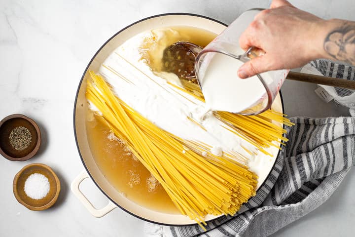A hand pouring half and half over a pot of noodles