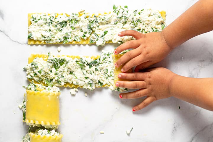 Two toddler hands rolling up cheesy spinach lasagna