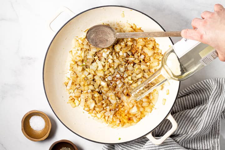 White wine being poured into a large white pot with sauteed onion and garlic