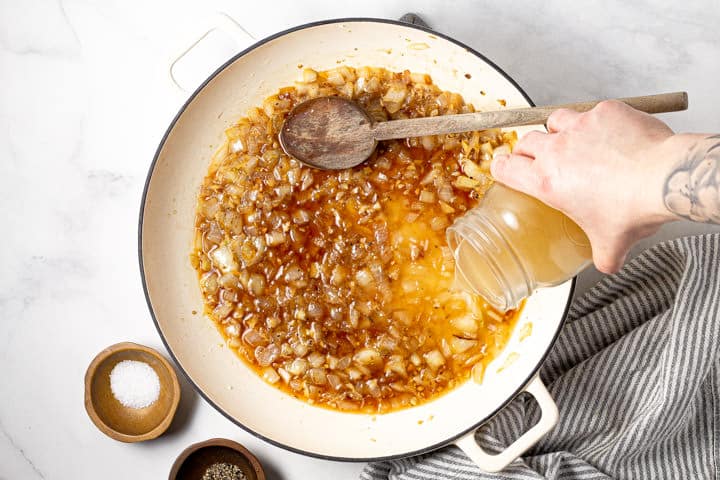 Veggie stock being poured into a large white pot filled with sauteed onions