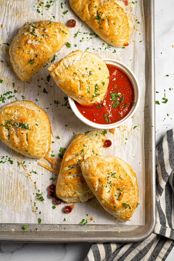 Overhead shot of baked mini calzones on parchment lined baking sheet with one of them being dunked in a bowl of pizza sauce 