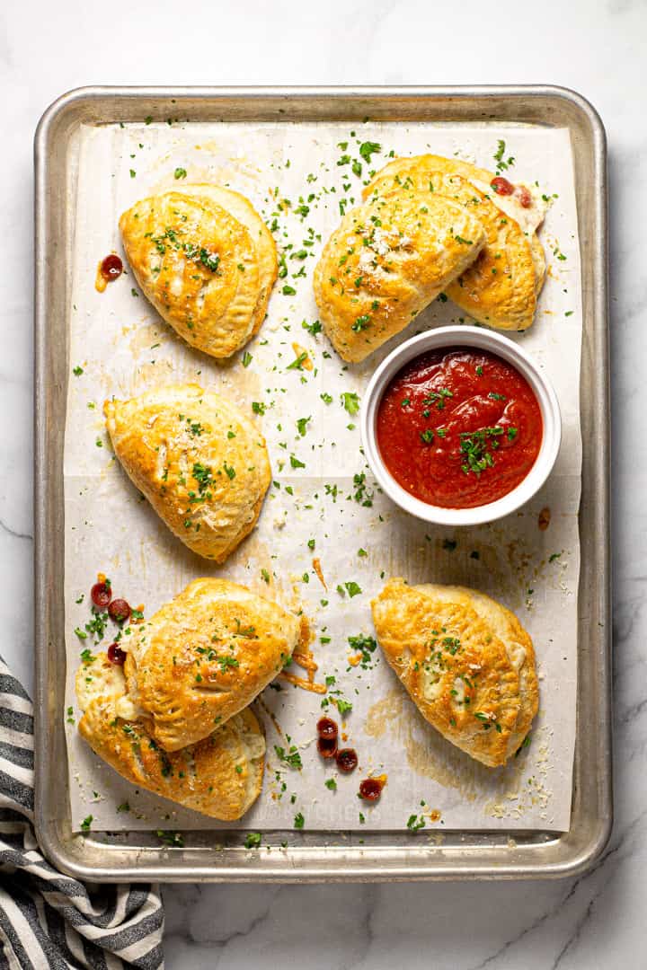 Parchment lined baking sheet with fresh baked mini calzones with a small bowl of pizza sauce 