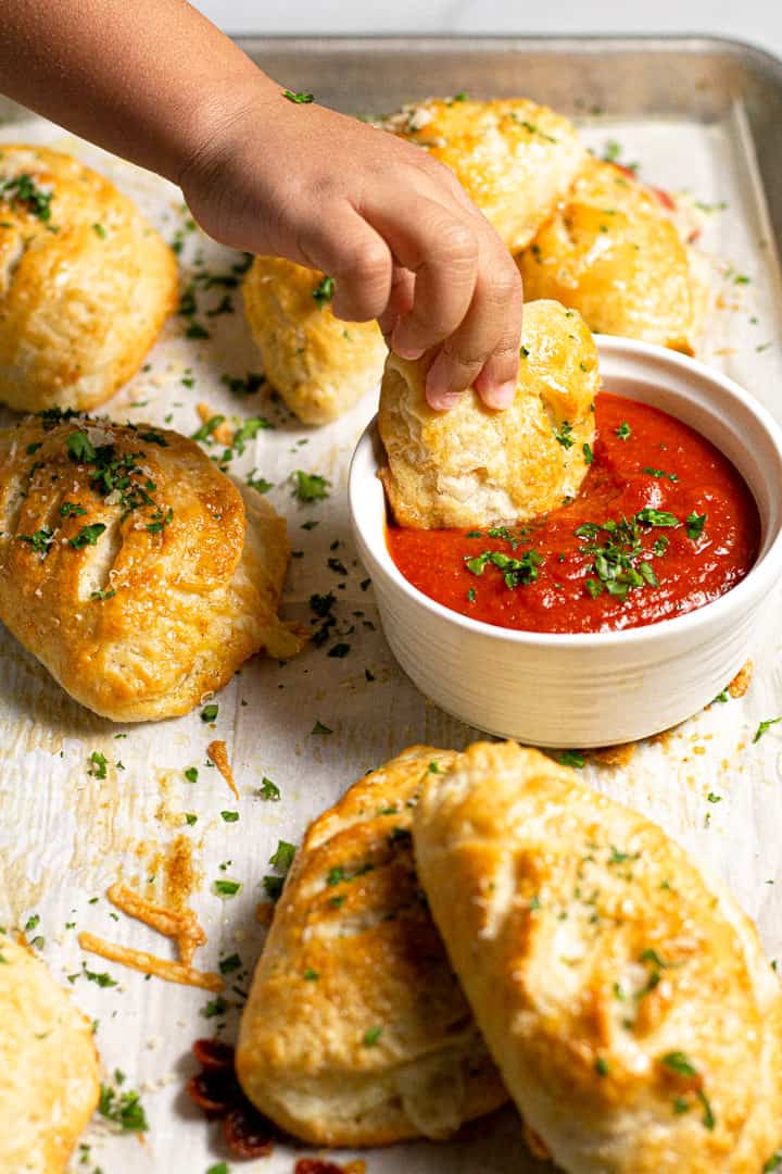 A small hand dunking a fresh baked mini calzone into a small bowl of pizza sauce 
