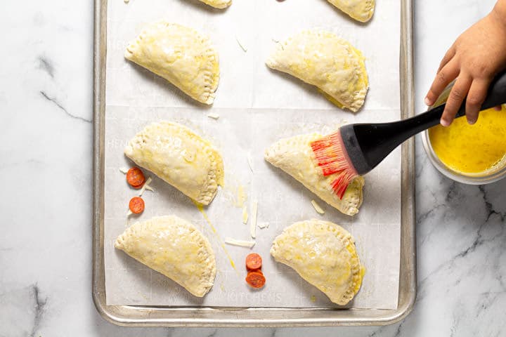 A small hand using a pastry brush to spread egg wash over unbaked calzones 