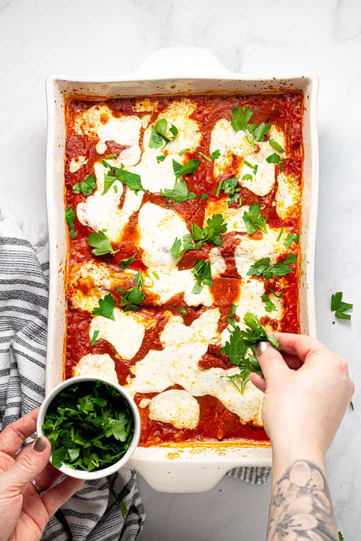 A white 9x13 pan of freshly baked manicotti being garnished with chopped fresh parsley
