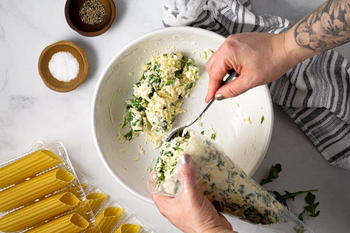 Two hands spooning manicotti filling into a ziploc baggy