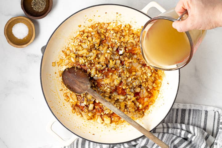Chicken broth being poured into a large white pot of sauteed onions