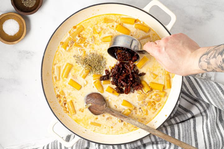 Sun dried tomatoes being added to a large white pot of pasta