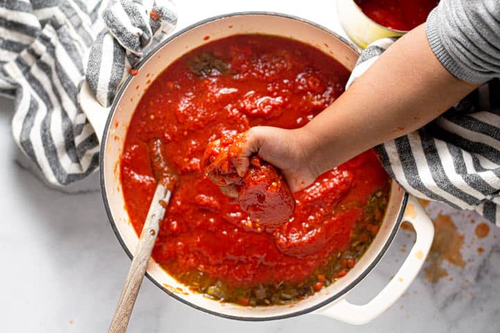 Small hand squeezing a whole San Marzano tomato over a pot of spaghetti sauce 