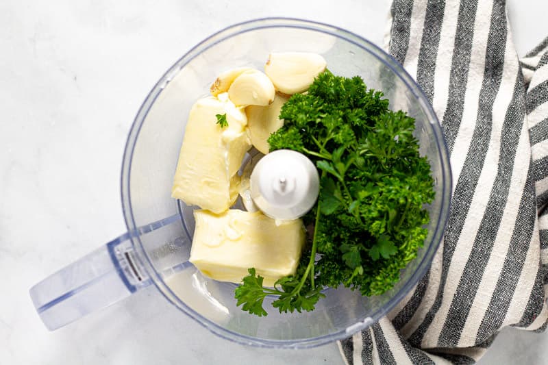 A food processor filled with softened butter garlic cloves and fresh parsley 
