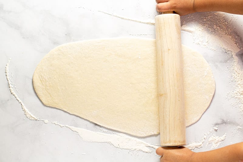 Fresh flatbread dough being rolled out on a white marble counter top 