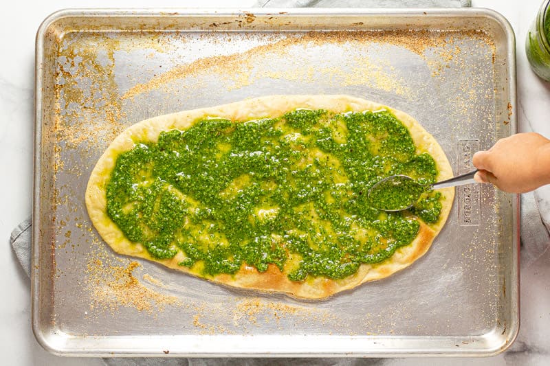 A spoon spreading fresh spinach pesto onto a flatbread crust on a baking sheet