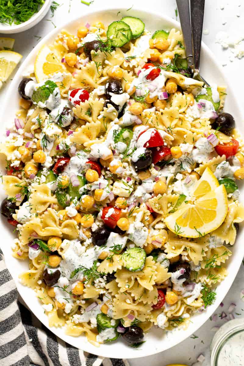 Overhead shot of a large white platter of Greek pasta salad drizzled with yogurt sauce