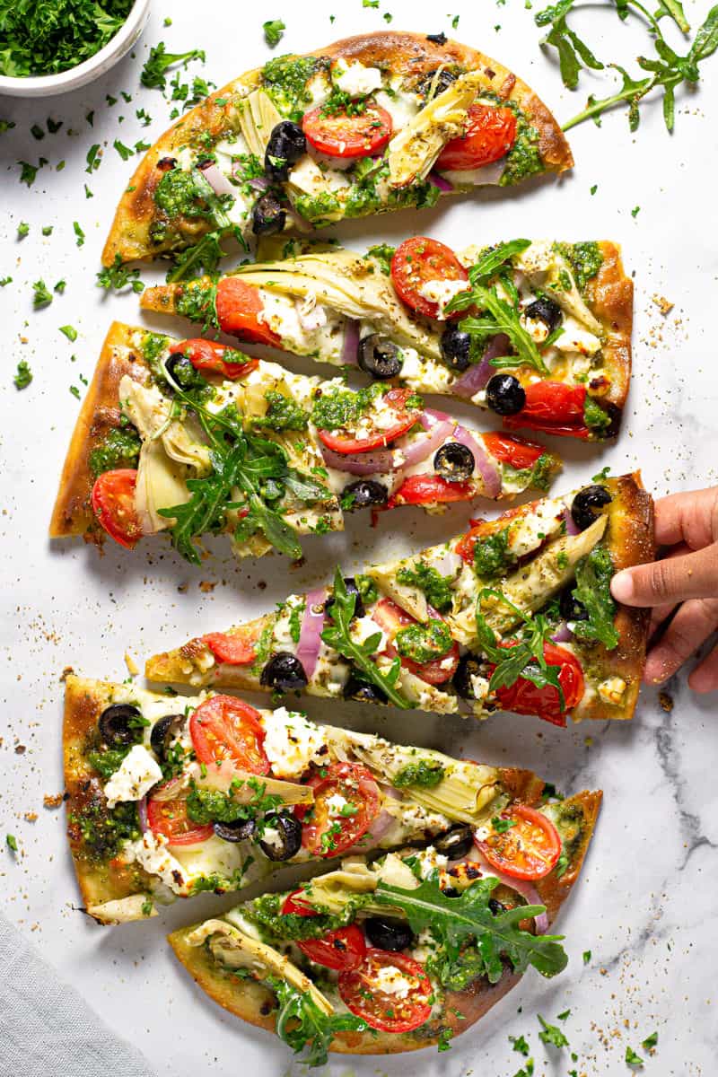 Sliced homemade Mediterranean veggie flatbread with a small hand reaching for a slice