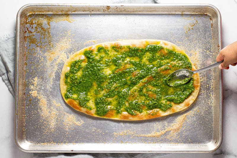 A small hand using a spoon to spread spinach pesto on a flatbread crust 