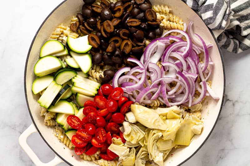 Zucchini olives red onion tomato and artichoke hearts being added to a pan of pasta