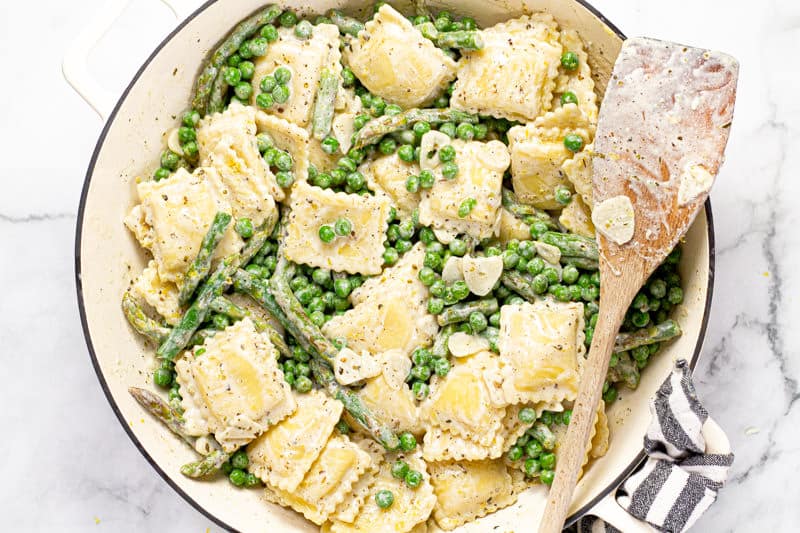 Large white pan with a creamy herb and garlic goat cheese sauce with ravioli peas and asparagus