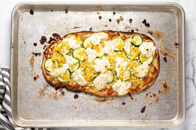 Baking sheet with a corn zucchini and ricotta flatbread on it