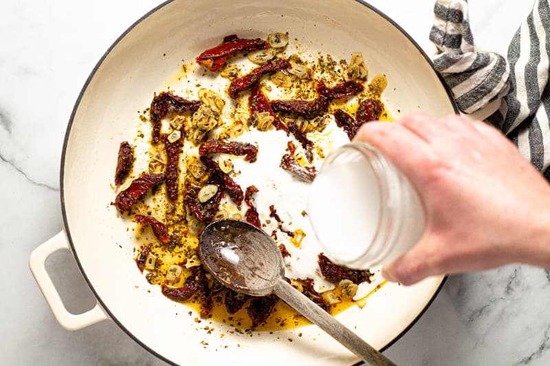 A hand pouring coconut milk into a pan of sun dried tomatoes and garlic