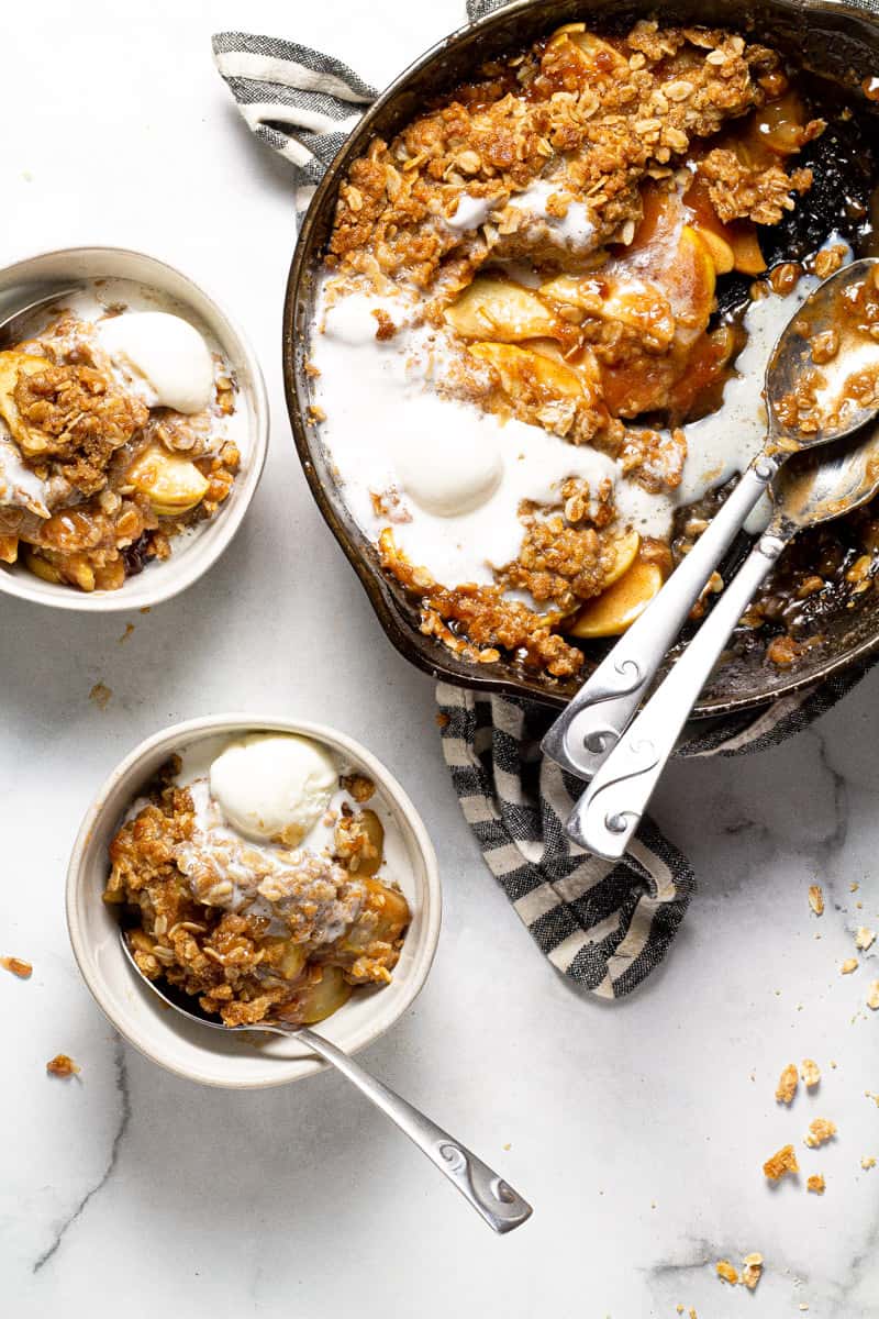 Two white bowls and a large cast iron skillet with apple crisp and vanilla ice cream 