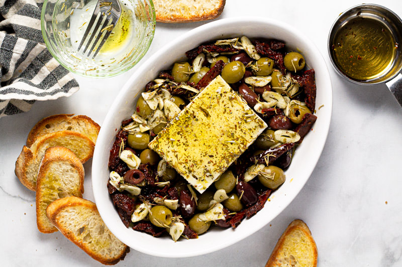 Small white baking dish with olives, sun dried tomatoes and a block of feta in it