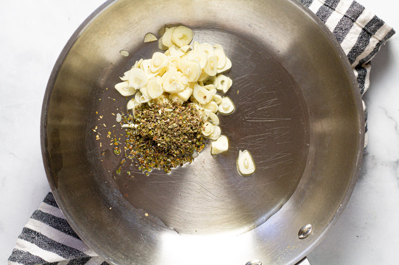 Metal pan with olive oil sliced garlic and dried oregano in it 