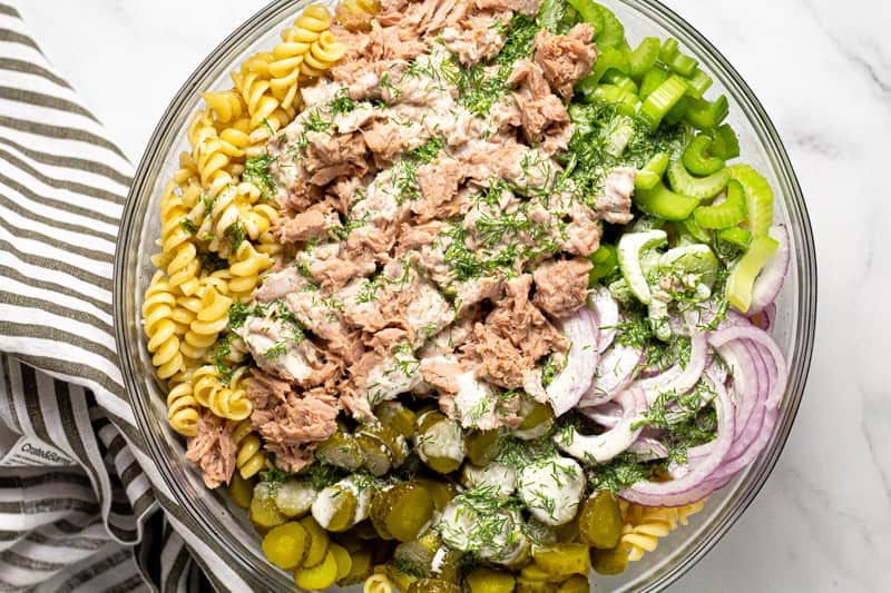 Large glass bowl with ingredients to make tuna pasta salad drizzled with homemade dressing