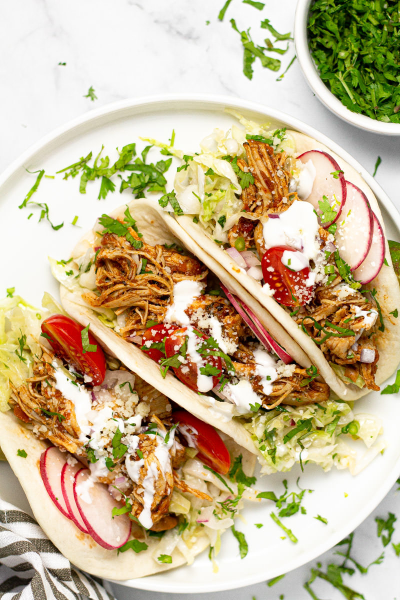 White plate with three shredded chicken tacos loaded with slaw and garnished with cilantro