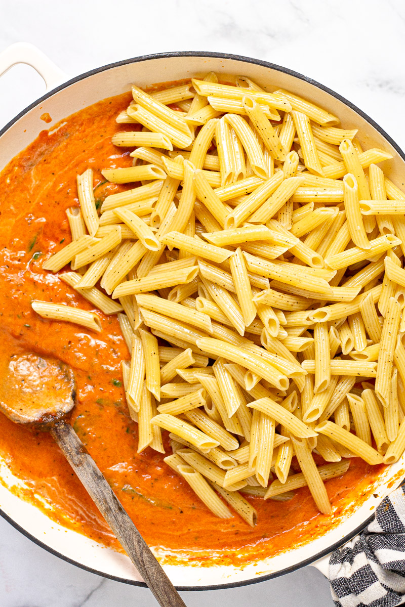 Al dente penne pasta being added to a tomato cream sauce in a large white pan 