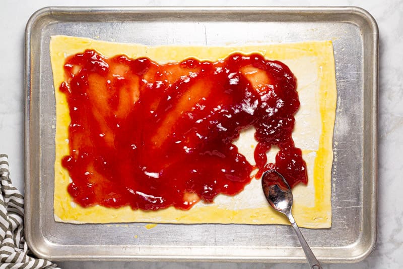 Baking sheet with puff pastry being spread with strawberry jam 