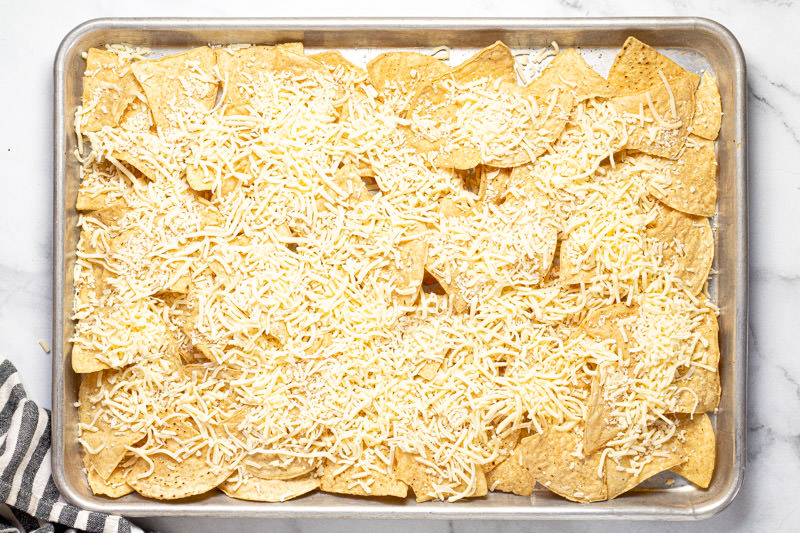Baking sheet pan with tortilla chips and shredded cheese spread in an even layer on it 