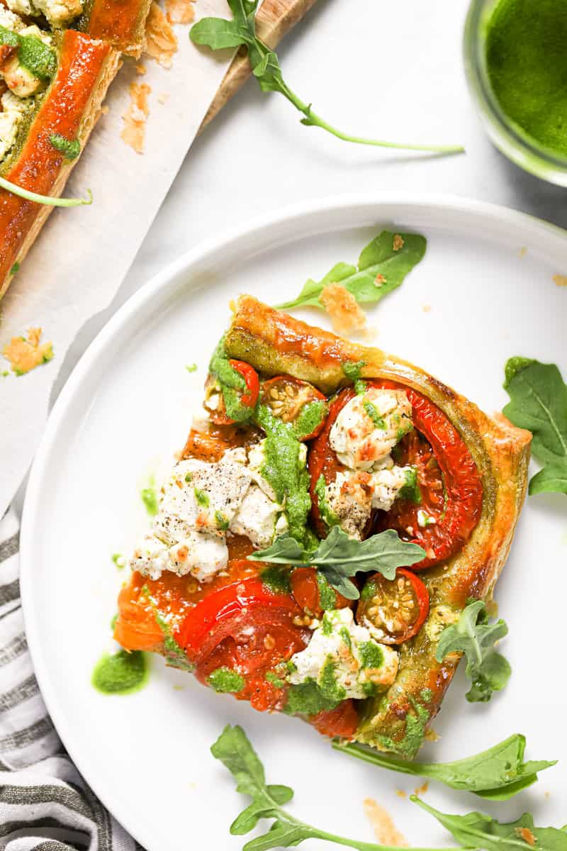 White plate with a slice of puff pastry tomato tart garnished with arugula