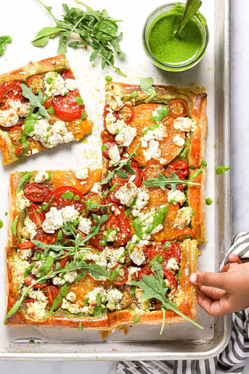 A small hand grabbing a slice of puff pastry tomato tart from a sheet pan