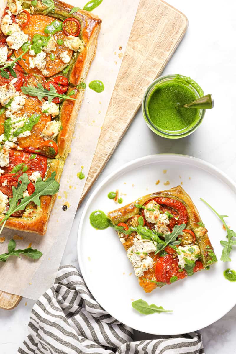 A slice of puff pastry tomato tart on a white plate next to a cutting board with remaining tart