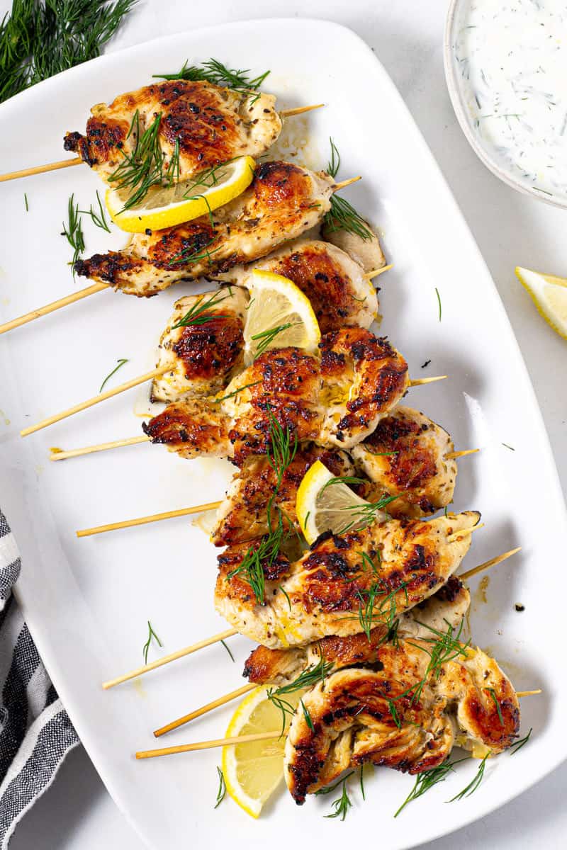 Overhead shot of chicken kebabs on a white platter garnished with fresh dill and lemon slices