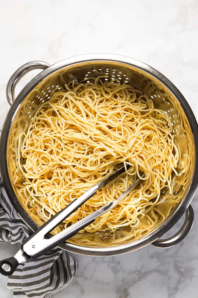 Cooked spaghetti in a metal strainer with metal tongs