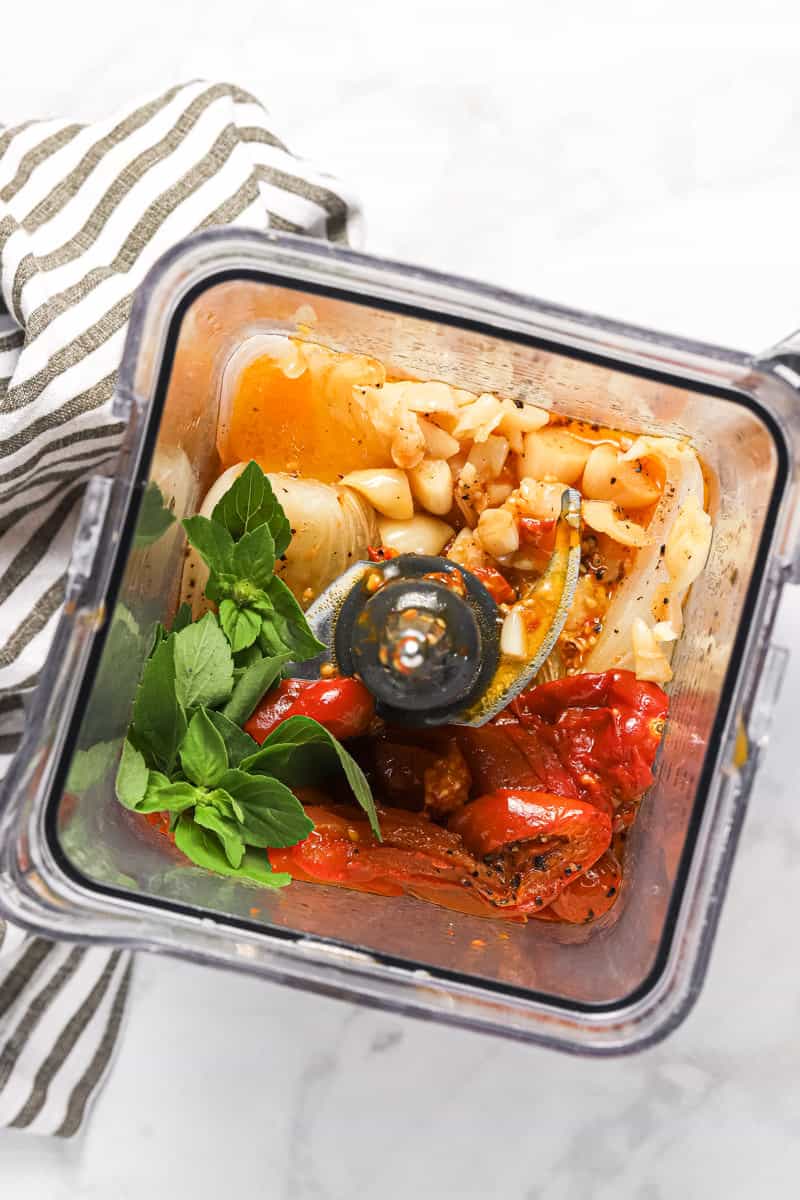 A blender filled with roasted veggies and fresh basil leaves