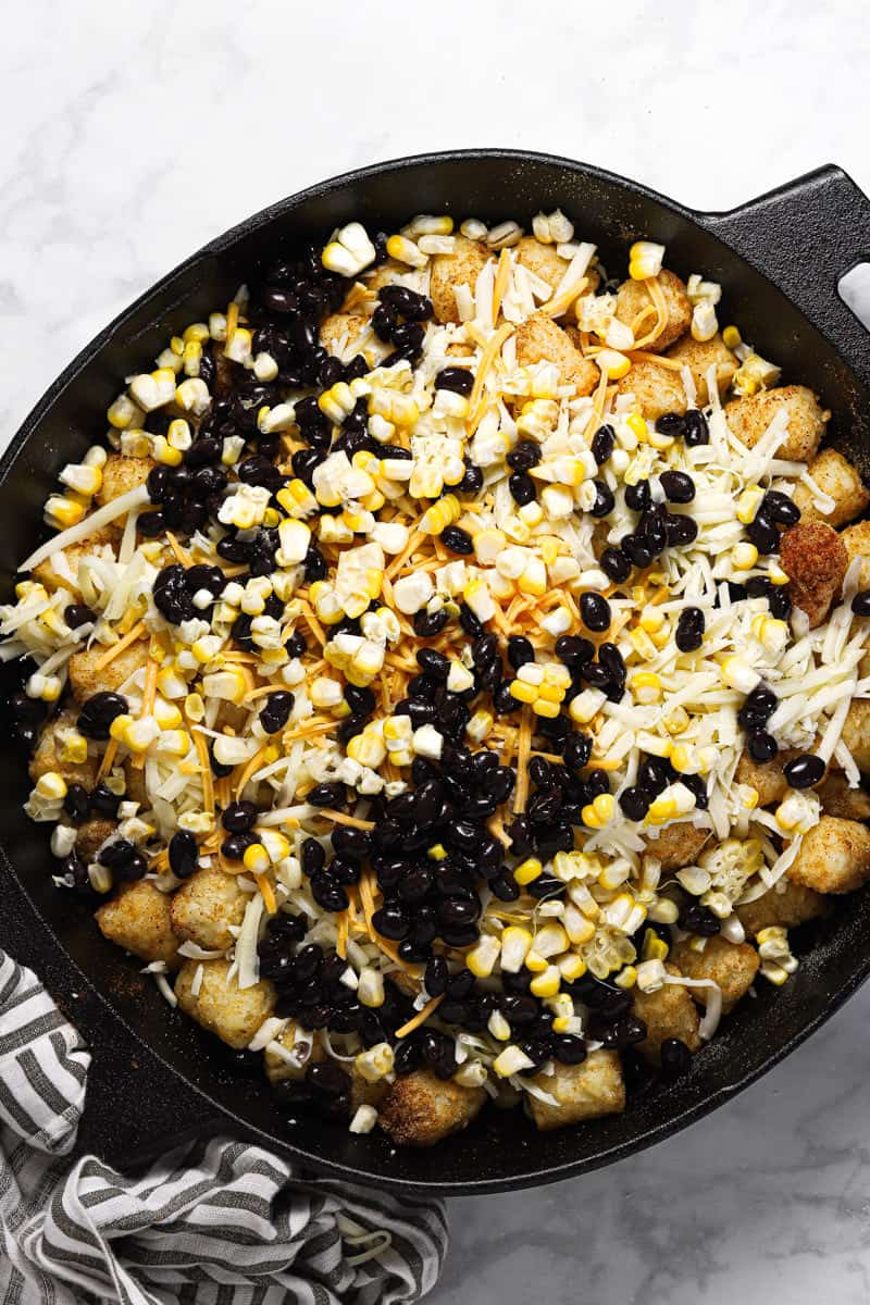 Tater tots in a cast iron skillet topped with cheese black beans and corn