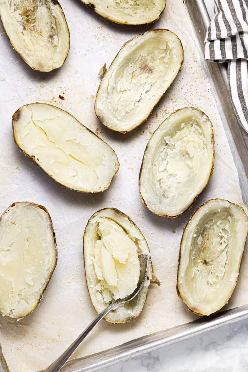 Baking sheet with halved baked potatoes with their centers being scooped out with a spoon